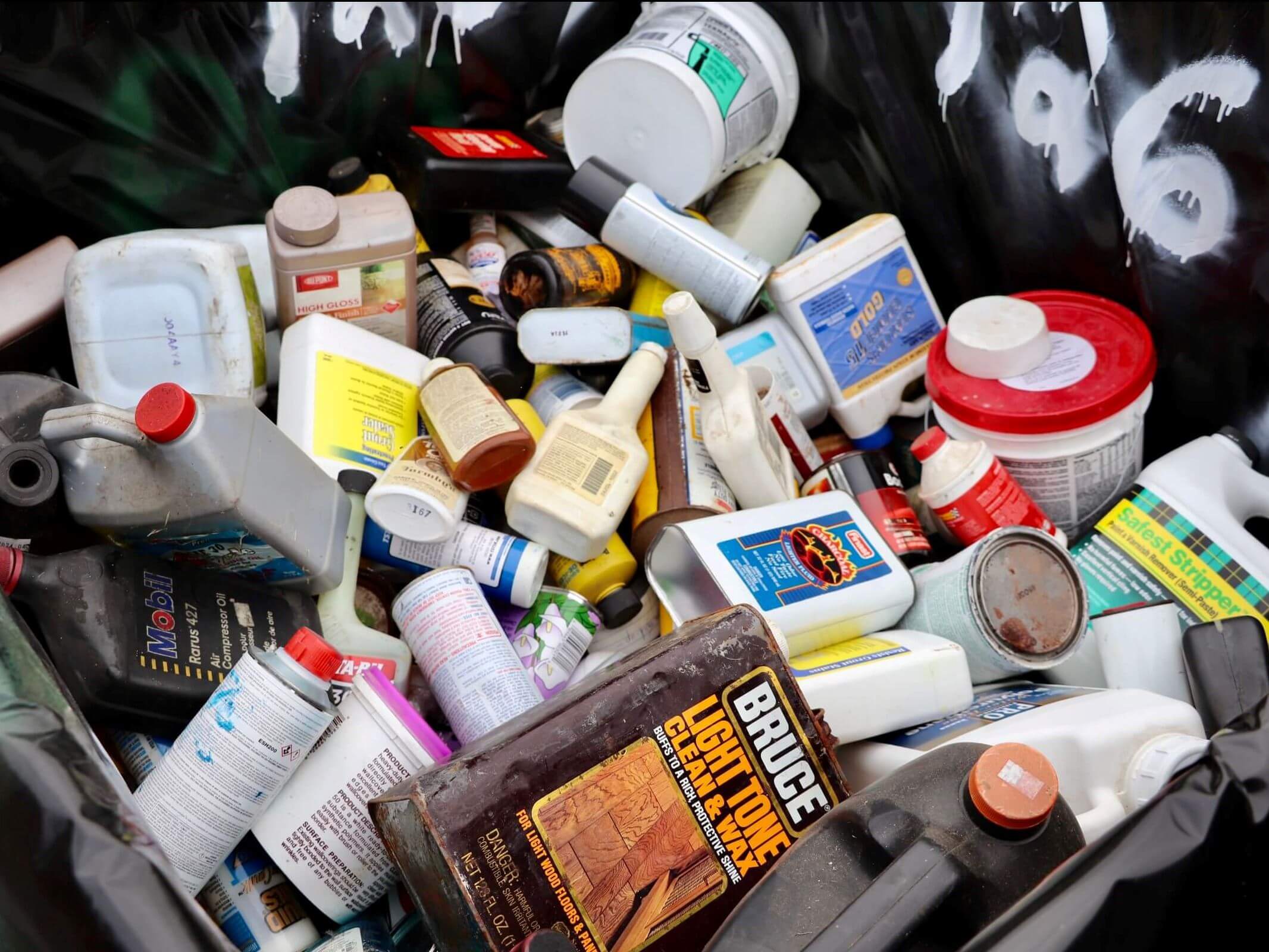 Collection of hazardous household waste including chemical bottles, cleaning products, paint cans, and various containers with red and white caps arranged on a dark surface. The items represent typical household hazardous materials that require proper disposal in compliance with environmental and safety regulations.